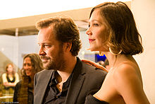 A man and a woman pose together for a photo. The man has short, light-brown hair and a beard, and is wearing a grey suit jacket and grey shirt. The woman has short, shoulder-length brown hair, worn loose, and is wearing large hoop earrings with a sleeveless, strapless black dress.